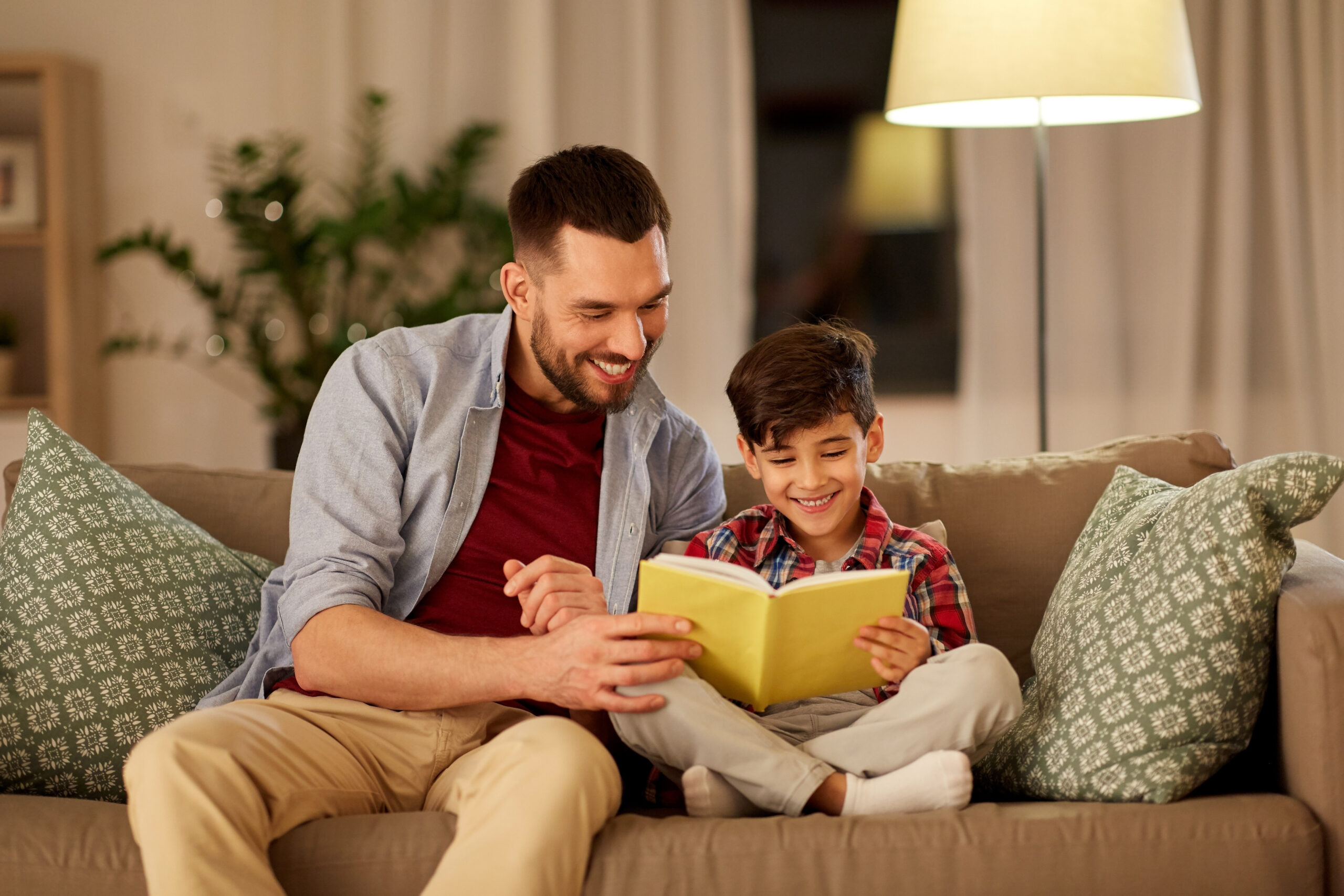 family, childhood, fatherhood, leisure and people concept - happy smiling father and little son reading book on sofa at home in evening