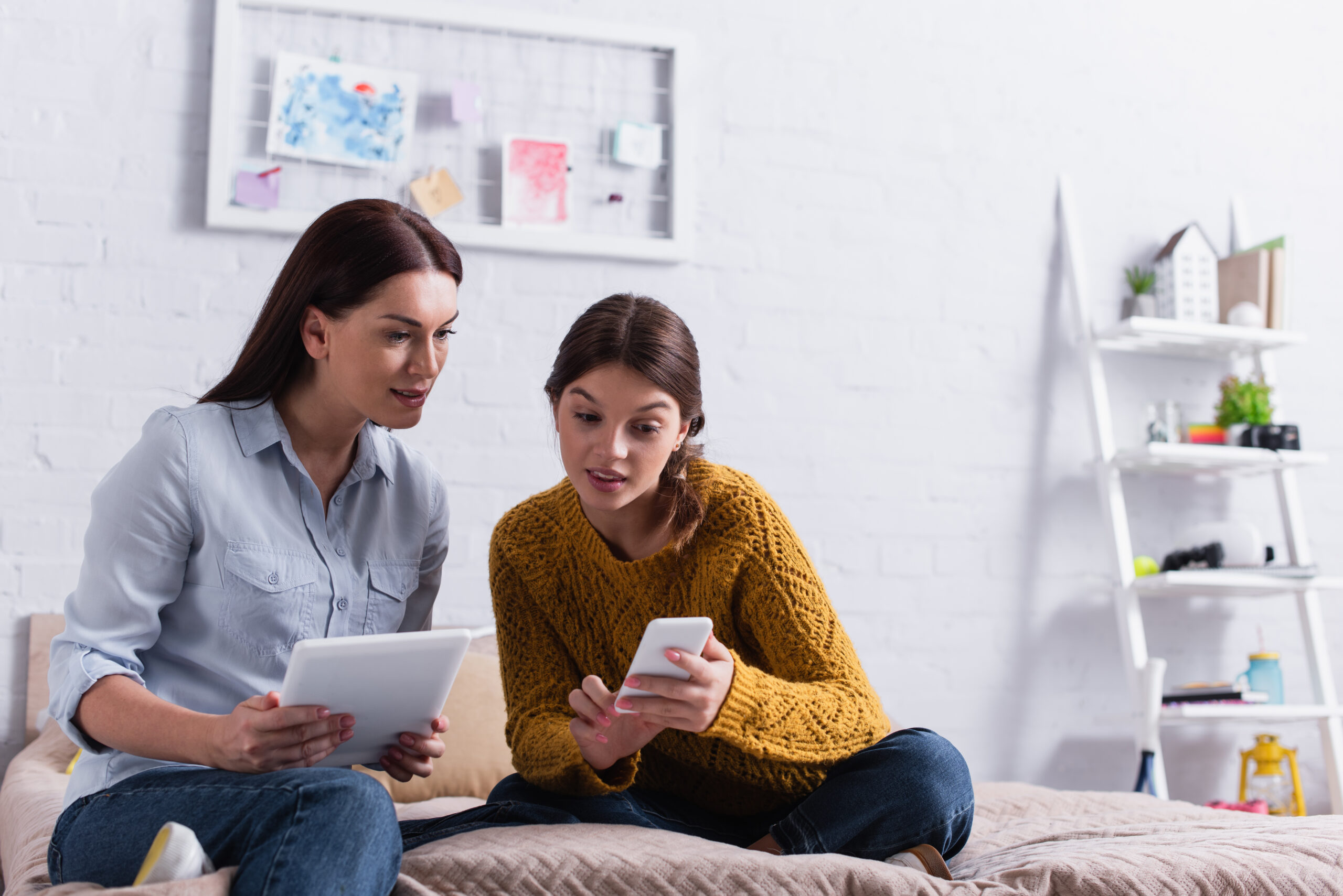 teenage girl holding smartphone near mother with digital tablet on bed