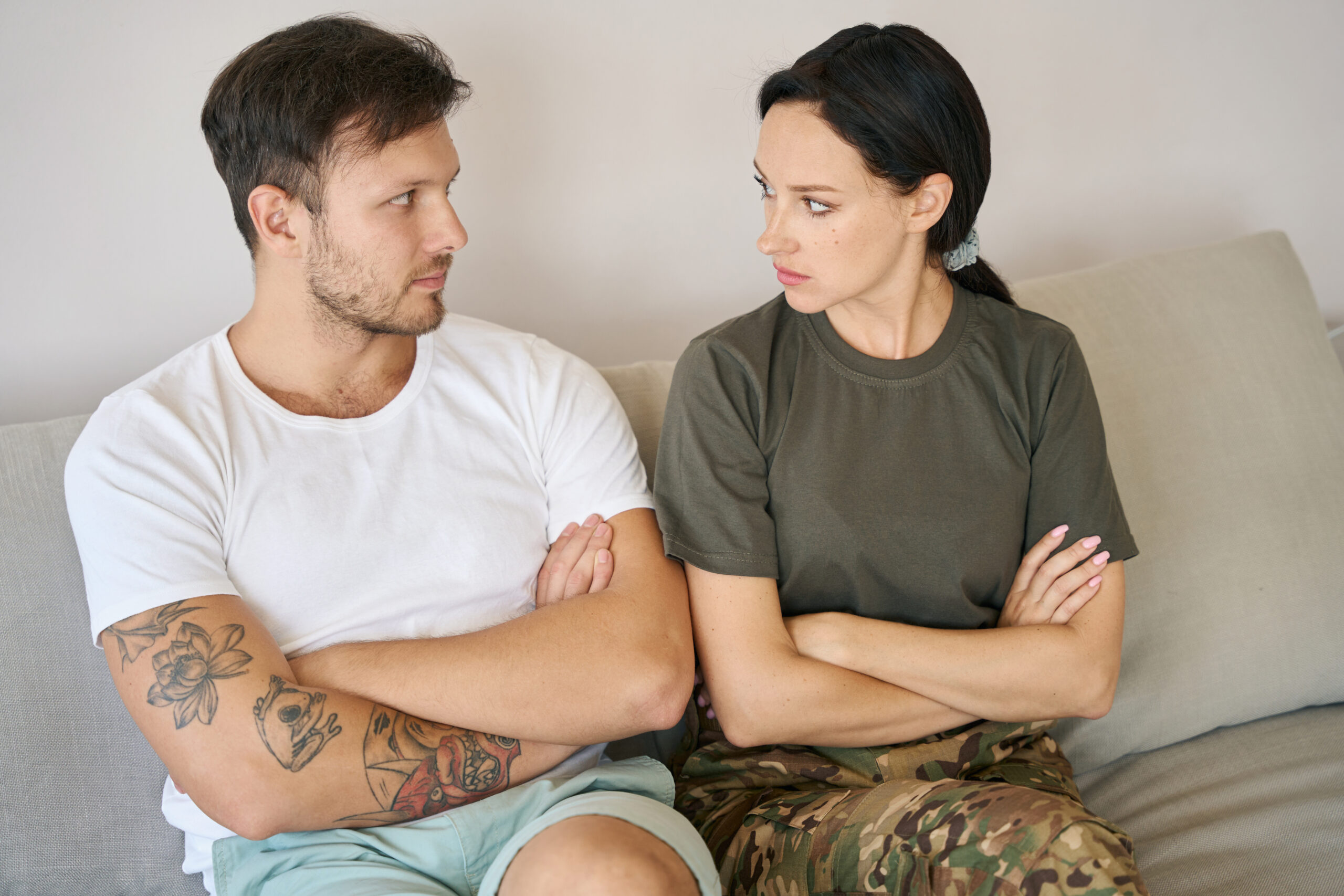 Young man and woman sitting side by side on couch in closed poses, they intensely looking into each other eyes